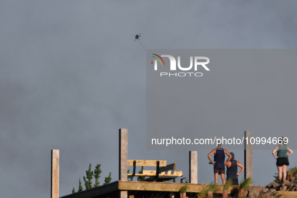 A fire-fighting helicopter is flying over an area of burning vegetation, working to extinguish a wildfire in Christchurch, New Zealand, on F...