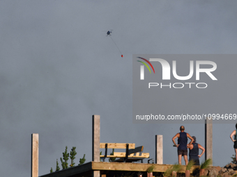 A fire-fighting helicopter is flying over an area of burning vegetation, working to extinguish a wildfire in Christchurch, New Zealand, on F...