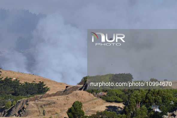 A fire-fighting helicopter is flying over an area of burning vegetation, working to extinguish a wildfire in Christchurch, New Zealand, on F...