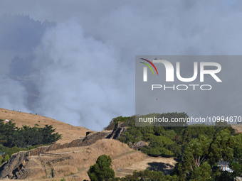 A fire-fighting helicopter is flying over an area of burning vegetation, working to extinguish a wildfire in Christchurch, New Zealand, on F...