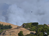 A fire-fighting helicopter is flying over an area of burning vegetation, working to extinguish a wildfire in Christchurch, New Zealand, on F...