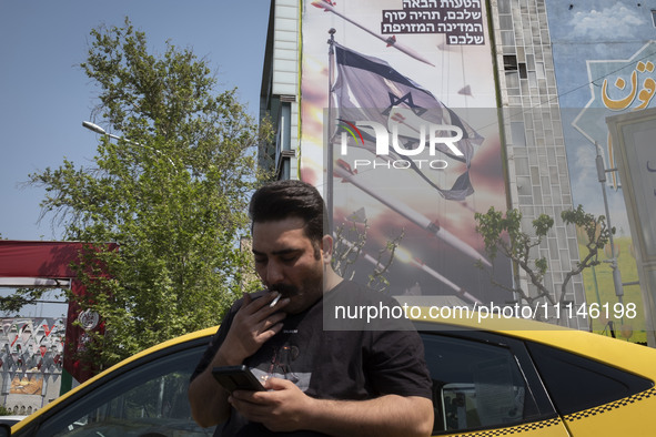 An Iranian yellow cab driver is using his cellphone while standing under an anti-Israeli mural, which appreciates the IRGC missile and UAV a...