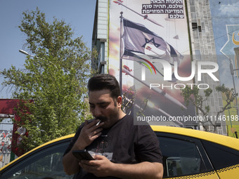 An Iranian yellow cab driver is using his cellphone while standing under an anti-Israeli mural, which appreciates the IRGC missile and UAV a...