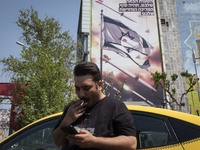 An Iranian yellow cab driver is using his cellphone while standing under an anti-Israeli mural, which appreciates the IRGC missile and UAV a...