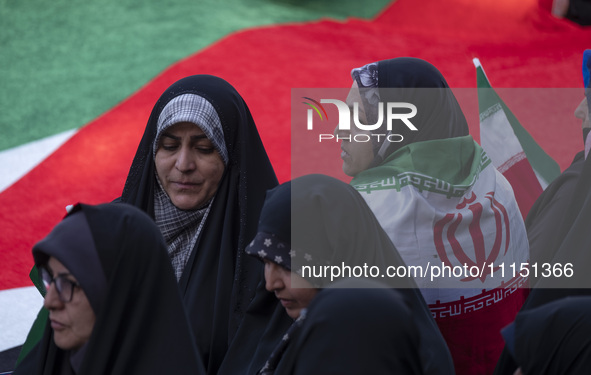 A veiled woman is standing next to a massive Palestinian flag while wearing an Iranian flag during a gathering to celebrate the IRGC UAV and...