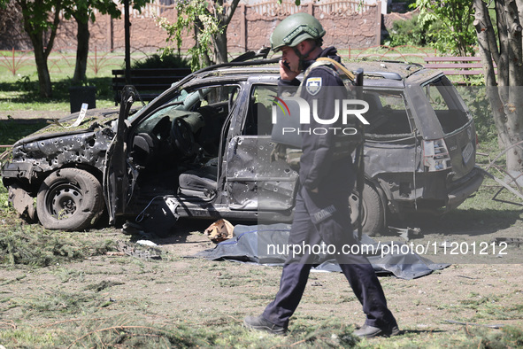 A police officer is talking on the phone while walking past the body of a victim in a human remains pouch lying on the ground by a damaged c...