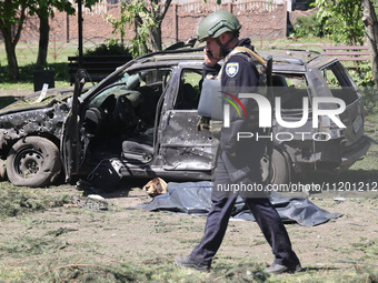 A police officer is talking on the phone while walking past the body of a victim in a human remains pouch lying on the ground by a damaged c...