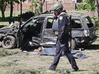 A police officer is talking on the phone while walking past the body of a victim in a human remains pouch lying on the ground by a damaged c...