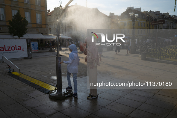 Tow women wearing headscarfs, hijabs are seen cooling off at a misting station in Warsaw, Poland on 28 June, 2024. 