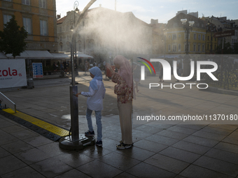 Tow women wearing headscarfs, hijabs are seen cooling off at a misting station in Warsaw, Poland on 28 June, 2024. (