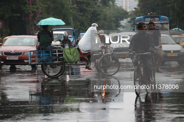 A man is sitting with an umbrella as another man is covering himself with a plastic sheet while paddling his rickshaw on a busy road during...