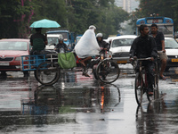 A man is sitting with an umbrella as another man is covering himself with a plastic sheet while paddling his rickshaw on a busy road during...