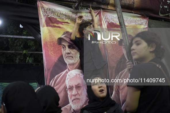 A young boy is standing in front of a banner featuring portraits of the former commander of the Hashd Shabi, Abu Mahdi al-Muhandes (bottom),...