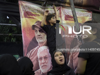 A young boy is standing in front of a banner featuring portraits of the former commander of the Hashd Shabi, Abu Mahdi al-Muhandes (bottom),...