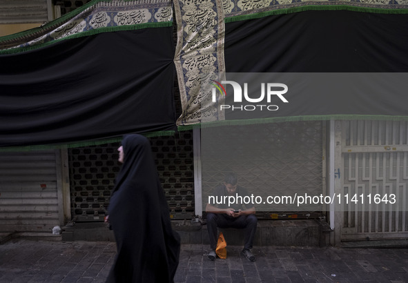 A young Iranian man is using his cellphone while sitting under a giant religious flag at a bazaar, before participating in a religious festi...