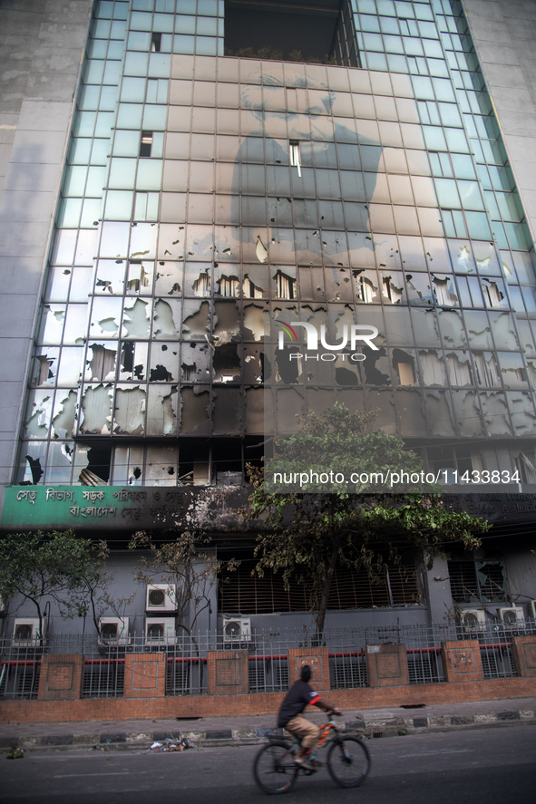 A cyclist is crossing past the burnt building of the Bangladesh Bridge Authority, which students are setting on fire amid the ongoing anti-q...