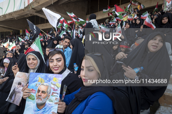 Two young Iranian women are sitting next to their veiled mother while holding a poster featuring a portrait of former commander of the Islam...