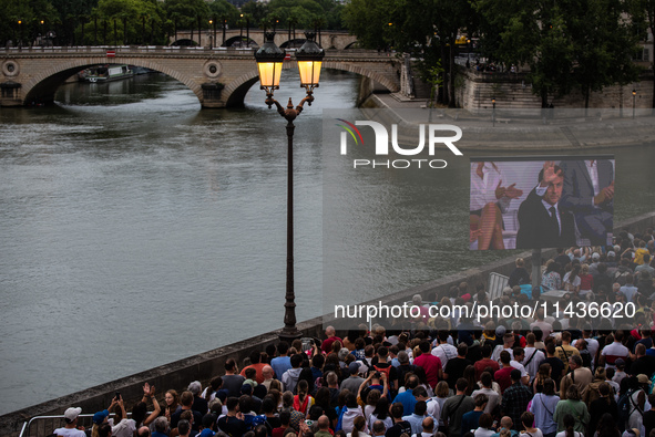 People are sitting on the benches of the Seine River, and Emmanuel Macron is appearing on screen during the opening ceremony of the Paris Ol...