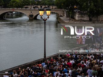 People are sitting on the benches of the Seine River, and Emmanuel Macron is appearing on screen during the opening ceremony of the Paris Ol...