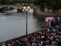 People are sitting on the benches of the Seine River, and Emmanuel Macron is appearing on screen during the opening ceremony of the Paris Ol...