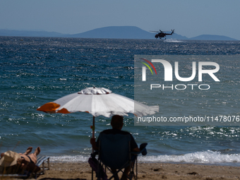 A swimmer is enjoying the sea while a firefighting helicopter is collecting water from it in Nea Makri, Greece, on August 14, 2024, as it is...