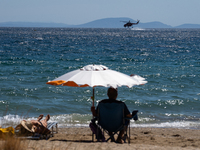 A swimmer is enjoying the sea while a firefighting helicopter is collecting water from it in Nea Makri, Greece, on August 14, 2024, as it is...