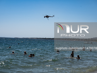 Swimmers are enjoying the sea while a firefighting helicopter is collecting water from it in Nea Makri, Greece, on August 14, 2024, as it is...