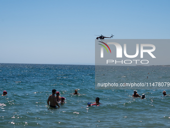 Swimmers are enjoying the sea while a firefighting helicopter is collecting water from it in Nea Makri, Greece, on August 14, 2024, as it is...