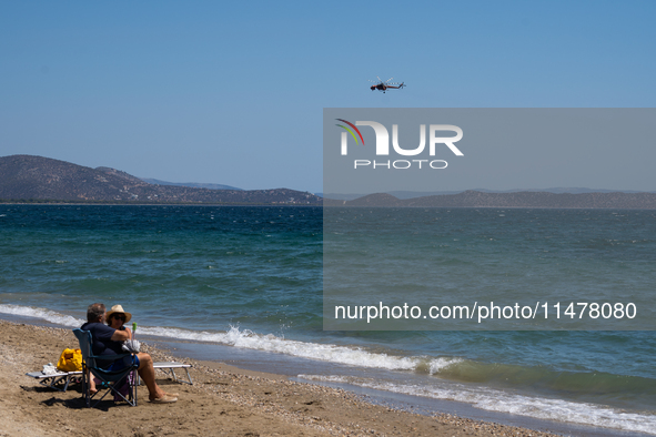 Swimmers are enjoying the sea while a firefighting helicopter is collecting water from it in Nea Makri, Greece, on August 14, 2024, as it is...