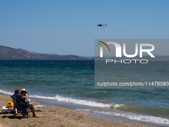 Swimmers are enjoying the sea while a firefighting helicopter is collecting water from it in Nea Makri, Greece, on August 14, 2024, as it is...