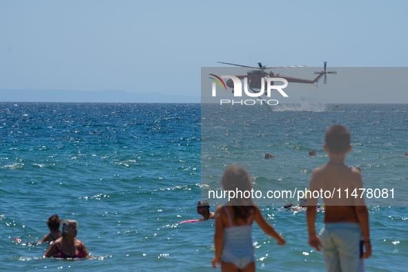 Two children are in the sea, watching the firefighting helicopter collect water from the sea in Nea Makri, Greece, on August 14, 2024, as it...