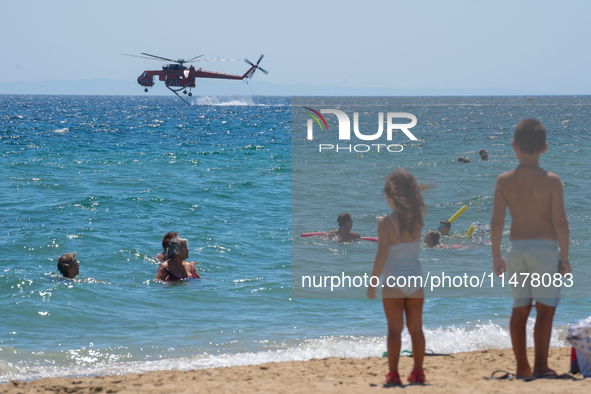 Two children are in the sea, watching the firefighting helicopter collect water from the sea in Nea Makri, Greece, on August 14, 2024, as it...