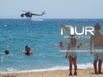 Two children are in the sea, watching the firefighting helicopter collect water from the sea in Nea Makri, Greece, on August 14, 2024, as it...