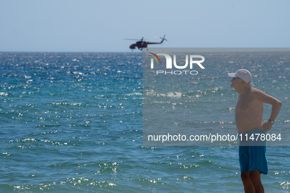 A swimmer is enjoying the sea while a firefighting helicopter is collecting water from it in Nea Makri, Greece, on August 14, 2024, as it is...