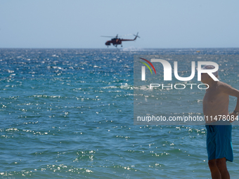 A swimmer is enjoying the sea while a firefighting helicopter is collecting water from it in Nea Makri, Greece, on August 14, 2024, as it is...