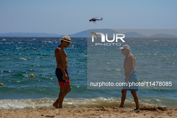 Bathers are enjoying the sea while a firefighting helicopter is collecting water from it in Nea Makri, Greece, on August 14, 2024, as it is...