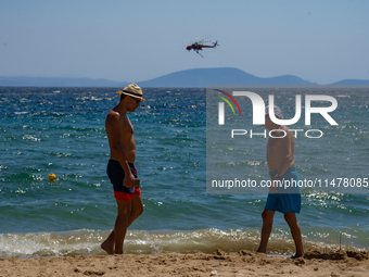 Bathers are enjoying the sea while a firefighting helicopter is collecting water from it in Nea Makri, Greece, on August 14, 2024, as it is...