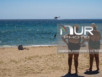 Bathers are enjoying the sea while a firefighting helicopter is collecting water from it in Nea Makri, Greece, on August 14, 2024, as it is...