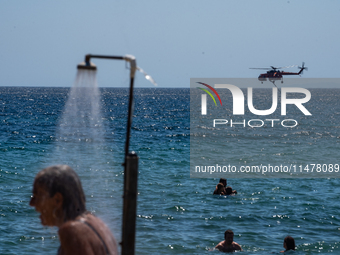 Bathers are enjoying the sea while a firefighting helicopter is collecting water from it in Nea Makri, Greece, on August 14, 2024, as it is...