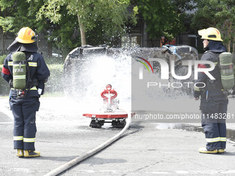 A firefighting robot is spraying water under the chassis of an electric vehicle that is simulated to be on fire and is lifting the vehicle t...