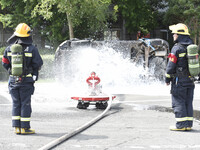 A firefighting robot is spraying water under the chassis of an electric vehicle that is simulated to be on fire and is lifting the vehicle t...