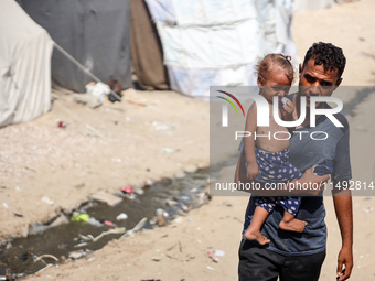 A displaced Palestinian father is carrying his son, who is suffering from a skin rash, while standing next to a pool of sewage water on the...