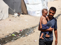 A displaced Palestinian father is carrying his son, who is suffering from a skin rash, while standing next to a pool of sewage water on the...