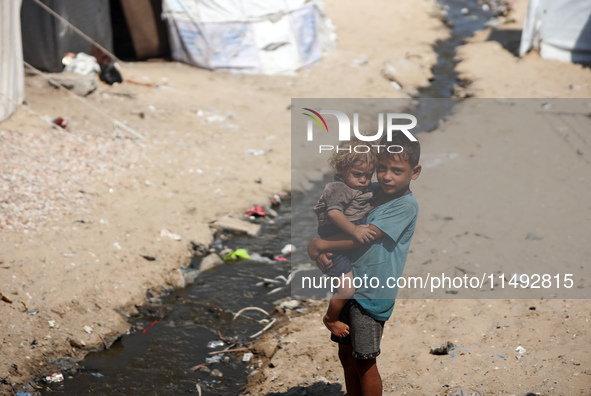 A displaced Palestinian boy is carrying his younger brother while standing next to their tent near the beach in Deir al-Balah in the central...