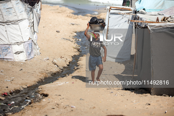 A displaced Palestinian boy is carrying kitchen utensils on his head and walking beside tents near the beach in Deir al-Balah in the central...