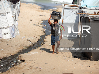 A displaced Palestinian boy is carrying kitchen utensils on his head and walking beside tents near the beach in Deir al-Balah in the central...