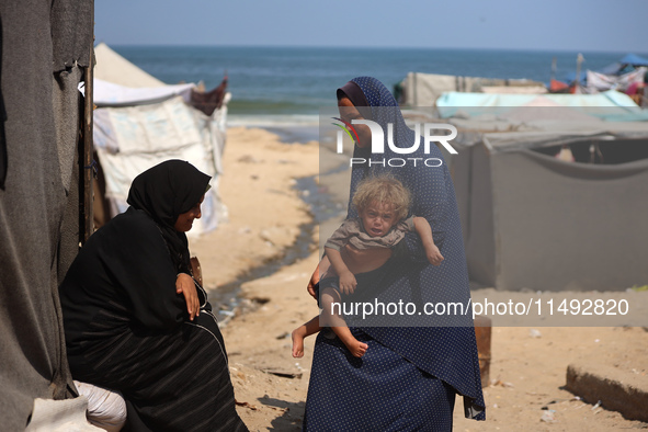 A displaced Palestinian mother is carrying her son, who is suffering from a skin rash, while standing next to a pool of sewage water on the...