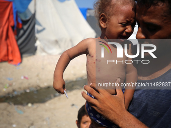 A displaced Palestinian father is carrying his son, who is suffering from a skin rash, while standing next to a pool of sewage water on the...