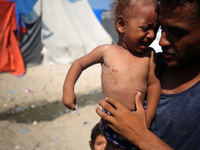 A displaced Palestinian father is carrying his son, who is suffering from a skin rash, while standing next to a pool of sewage water on the...