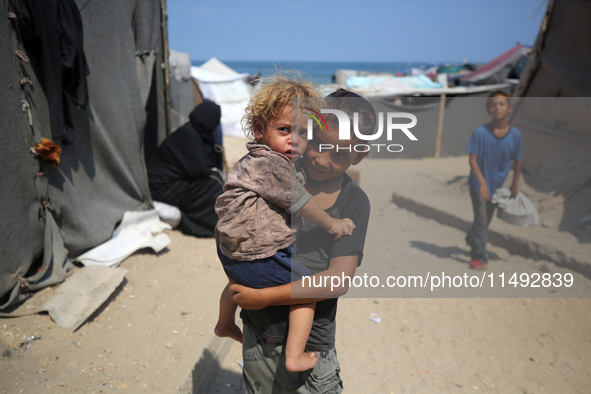 A displaced Palestinian boy is carrying his younger brother while standing next to their tent near the beach in Deir al-Balah in the central...